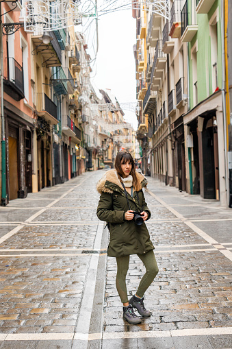 Young woman, brown hair, green coat, explores Pamplona's urban and rural fusion.