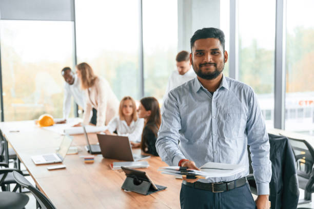 istock Indian man is standing in front of colleagues that are using laptop. Group of office workers are together indoors 1832442396
