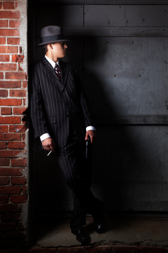 A man dressed in 1940s attire lurks in the shadows of a seedy warehouse district. He's holding a cigarette and a gun.
