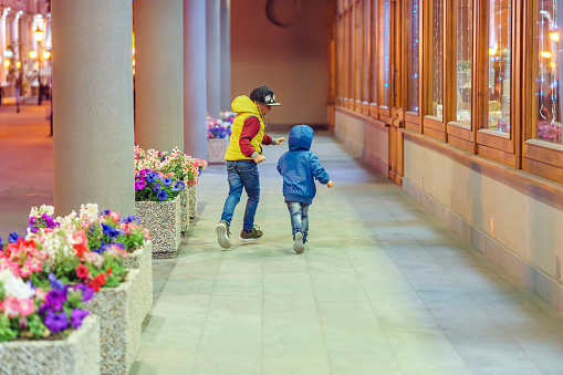 Happy little boys runs together along gallery between columns row with flowers and building facade at late evening