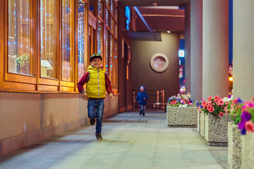 Happy little boys runs together along gallery between columns row with flowers and building facade at late evening
