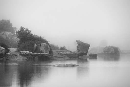 A black and white photograph featuring an idyllic landscape featuring a lake with rock formations appearing from the fog