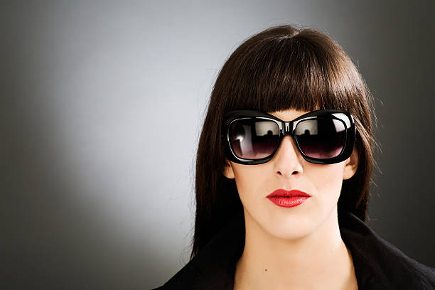 One highly sophisticated woman. She is looking straight at camera and wearing dark sunglasses and a black raincoat. Her lips are painted bright red.