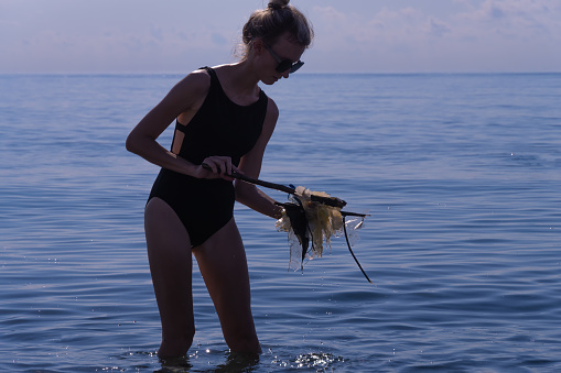 Young woman in a black one-piece swimsuit with the help of a stick collects floating cellophane bags right into the sea. Ecological catastrophe of the modern world, environmental pollution.