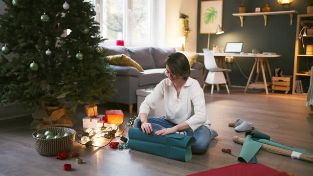 Young Caucasian woman wrapping Christmas present, while sitting near the Christmas tree