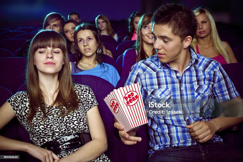 Teen couple in movie theater "Teenaged couple sitting in the cinema. The guy wants to give popcorn to the girl, but she seems to be dissatisfied. Audience in the background." Movie Theater Stock Photo