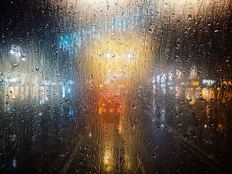 Abstract image depicting glowing city lights - coming from traffic and storefronts - defocused beyond a raindrop-soaked window.