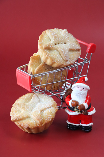 Stock photo showing figurine of Santa Claus standing besides a miniature shopping cart containing freshly baked, homemade, mince pies, against a red background. These individual mince pies are made with homemade short crust pastry glazed with an egg wash and sprinkled with sugar. The pies have been topped with pastry star detail, hiding the sweet mincemeat (mixture of chopped dried fruit, distilled spirits and spices) filling. Christmas festive food shopping concept.