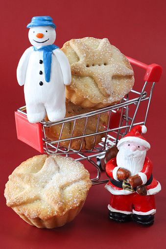 Stock photo showing figurine of Santa Claus standing besides a miniature shopping cart containing freshly baked, homemade, mince pies and a snowman model, against a red background. These individual mince pies are made with homemade short crust pastry glazed with an egg wash and sprinkled with sugar. The pies have been topped with pastry star detail, hiding the sweet mincemeat (mixture of chopped dried fruit, distilled spirits and spices) filling. Christmas festive food shopping concept.