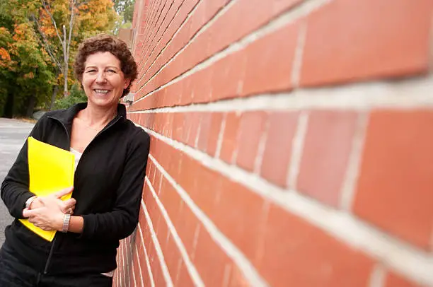 A mid-adult woman holding a yellow folder leans against a brick wall outside on a fall day.