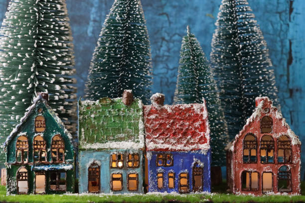 Image of scenic Christmas village display, artificial plastic spruce Christmas trees on artificial grass and snow of fleece surrounding row of illuminated homemade clay houses, focus on foreground
