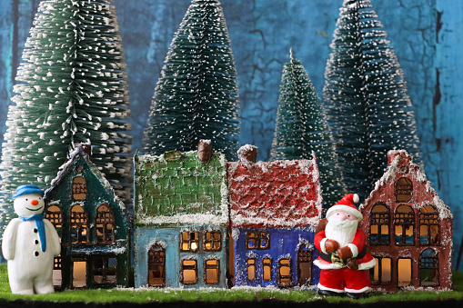 Stock photo showing a beautiful Christmas village display, which features numerous illuminated wooden houses. The houses have been placed on fake grass and snow to create a snowy mountain scene, in front of a blue backdrop, complete with a forest of plastic and wooden trees.