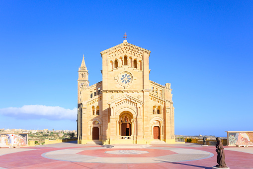 The Basilica of the National Shrine of the Blessed Virgin of Ta' Pinu is a Roman Catholic minor basilica and national shrine located near Garb on the island of Gozo, the sister island of Malta.\nIn 1883 a woman from the village of Garb, Karmni Grima, heard the voice of Our Lady at the small chapel that then occupied this site. It rapidly became a centre of pilgrimage and the number of visitors soon overwhelmed the little church.\nTodays magnificent church in Romanesque Style was built between 1920 and 1932. It is an architectural masterpiece, especially inside with its superb sculptures and craftsmanship in Maltese stone. The sanctuary was constructed in front of the original chapel. In 1932 the new Church was blessed and opened to the public and in 1935, Pope Pius XI raised it to the dignity of Minor Basilica.\nSince 2017 the space in front of basilica no longer serves as a carpark but with mosaic on both sides it became a place to welcome the devout apart from serving as an attraction and a spiritual aid to pilgrims. The mosaic work was commissioned to the Centro Aletti of Rome, under the guidance of Fr Marko Ivan Rupnik. The Centro Aletti is renowned for the sacred art which it has created in 150 churches all over the world, including the Vatican and sanctuaries such as Lourdes and Fatima.\nTa' Pinu, Gharb, Gozo, Malta, Europe.