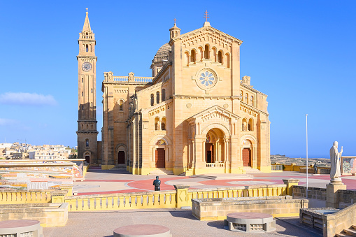 The Basilica of the National Shrine of the Blessed Virgin of Ta' Pinu is a Roman Catholic minor basilica and national shrine located near Garb on the island of Gozo, the sister island of Malta.\nIn 1883 a woman from the village of Garb, Karmni Grima, heard the voice of Our Lady at the small chapel that then occupied this site. It rapidly became a centre of pilgrimage and the number of visitors soon overwhelmed the little church.\nTodays magnificent church in Romanesque Style was built between 1920 and 1932. It is an architectural masterpiece, especially inside with its superb sculptures and craftsmanship in Maltese stone. The sanctuary was constructed in front of the original chapel. In 1932 the new Church was blessed and opened to the public and in 1935, Pope Pius XI raised it to the dignity of Minor Basilica.\nSince 2017 the space in front of basilica no longer serves as a carpark but with mosaic on both sides it became a place to welcome the devout apart from serving as an attraction and a spiritual aid to pilgrims. The mosaic work was commissioned to the Centro Aletti of Rome, under the guidance of Fr Marko Ivan Rupnik. The Centro Aletti is renowned for the sacred art which it has created in 150 churches all over the world, including the Vatican and sanctuaries such as Lourdes and Fatima.\nTa' Pinu, Gharb, Gozo, Malta, Europe.
