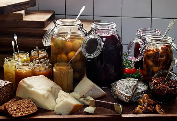 Still life with glass jars filled with homemade preserves, jars of jam and mustard,cheeses and freshly bread.