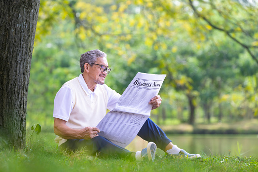 Senior Asian man reading a business newspaper while sitting under the tree by the lake at public park for recreation, leisure and relaxation in nature