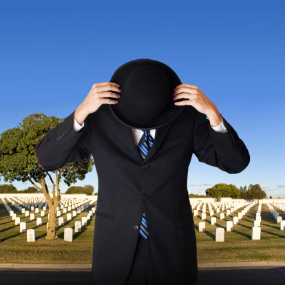 A man in a suit hides his head in his bowler hat. Cemetery in the background.Grave names are unreadable but appear to be normal names.