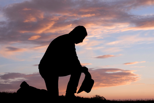 Silhouette of cowboy kneeling and holding hat with head bowed in prayer.