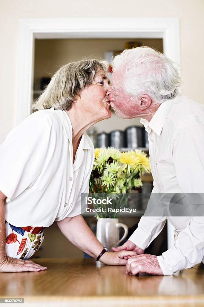 Sweet elderly couple hold hands and kiss Evidence of  a lasting romance as this elderly couple hold hands and kiss over their dining table. 70-79 Years Stock Photo