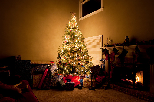 A brightly lit Christmas Tree in a dark living room on Christmas Eve with presents wrapped and piled up all around the tree.  A fire place burns with stocking hung with care.