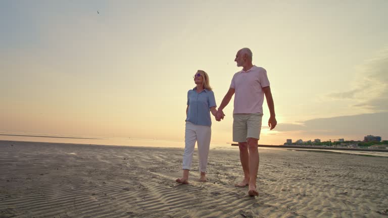 SLO MO TS Senior couple walking barefoot on a sandy beach at sunset and holding hands