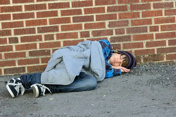 Runaway teenage boy sleeping against a brick wall in an alley.