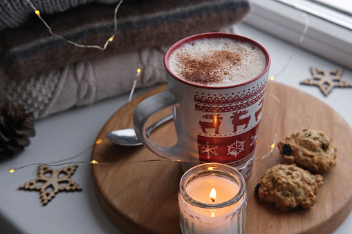 Winter windowsill still life. Red ceramic cup of hot coffee on window sill. Christmas decorations on the background. Cozy home picture. Warm woolen knitted sweaters, Burn Candle, Cookies. Stock photo. Winter still life. Calm cozy winter scene.