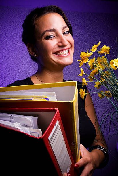 A young Caucasian woman with long blonde hair stands behind an enormous, teetering stack of papers and files that rests on a dark wooden desk. She is wearing a casual blue button-down shirt with rolled-up sleeves. She holds a pen in her right hand and is writing or signing a document on the very top of the pile, which is nearly as tall as she is. Her left hand rests on her forehead in a gesture of stress, exhaustion, or deep concentration. The background features a room with yellow walls and a white door, suggesting a home office or workplace environment. This image illustrates concepts of bureaucracy, excessive workload, tax season preparation, administrative backlog, or cramming for academic exams. The sheer volume of paperwork emphasizes the feeling of being overwhelmed by tasks and deadlines.