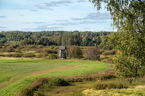 Landscape with a green meadow and a path leading to an old windmill against the background of a forest on a bright sunny autumn day.