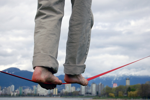 A young man delicately balances as he walks along a slackline (like a tightrope) with the cityscape of Vancouver, British Columbia and its classically cloudy sky in the background.