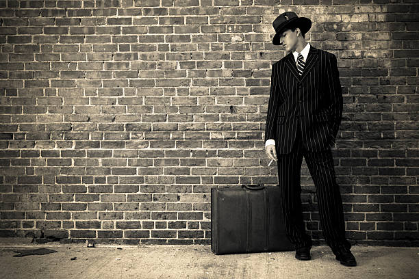 A young man dressed in 1940s attire standing in front of a brick wall with a suitcase.