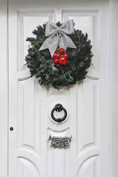 White front door decorated with festive Christmas wreath, made of lush green branches, is embellished with large silver bow and glossy red baubles. And decorative sign with the words MERRY XMAS