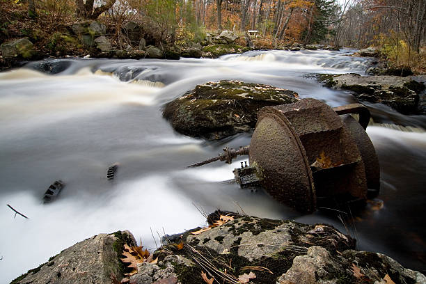 Picture of a rusting water wheel that has fallen into a stream bed.  Some of the original gears are still attached and can be see in the water.