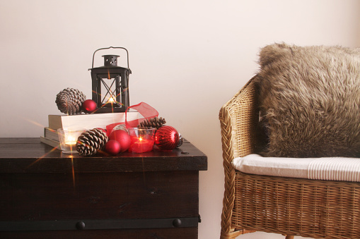 A studio shot of a home decor scene in winter time or Christmas. A wicker chair with warm and cozy furry pillow stands next to a wooden trunk table decorated with latern, books, candles, pine cones and Christmas ornaments.