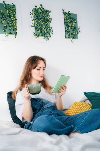 istock Young woman reading e-book on tablet reader in bed. Female relaxing with hot drink while reading an electronic book in cozy room. Mental health recovery, Personal growth. Hobby, Leisure time alone. 1832826195
