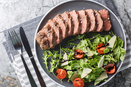 Sliced Seared Steak Tagliata di Manzo served with arugula, cherry tomatoes and parmesan close-up in a plate on the table. Horizontal top view from above