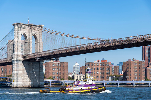 New York City, NY, USA-October 10, 2023; Suspension tower of the Brooklyn bridge over the East River with the lower eastside of Manhattan in the background and colorful tugboat on water