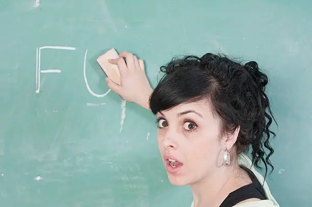A teacher erases profanity from a blackboard in a school classroom.  Teacher is a young woman next to the black board.  She is shocked.  Good copy space.
