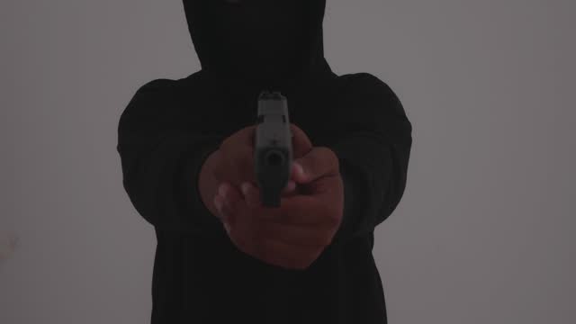 Mysterious man wearing black hoodie holding a pistol wearing a cloth mask covering his face completely in front of the door, Silhouette and dark concept image,threat of firearms,attack or defense.