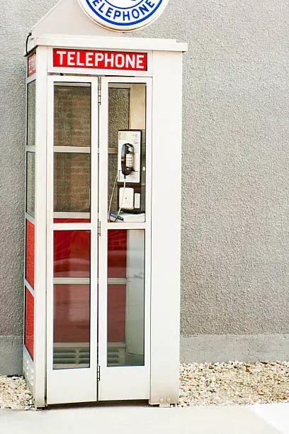 "Subject: Vertical view of a classic, old-fashioned American phone booth next to a stucco building wall."