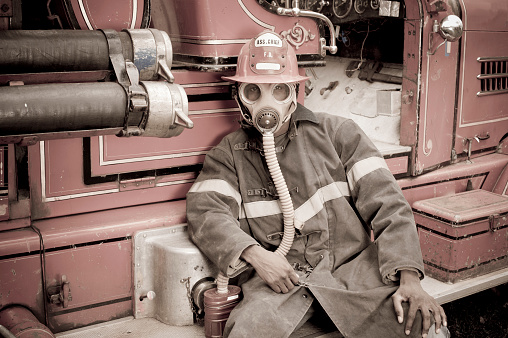A fireman or firefighter sits on a 1930s antique fire engine truck in authentic pre 1950 clothing and equipment.  Colour processed to look aged like an old photograph.  Click to view similar images.