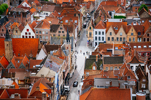 An aerial view of a Bruges town with red roofs. The buildings are tightly packed together and the streets are narrow. The photo is taken from a high angle, looking down on the town. The buildings are mostly made of brick and have a variety of architectural styles. The roofs are mostly red and orange, with some gray and white mixed in. The photo is taken during the day, with the sun shining on the buildings.