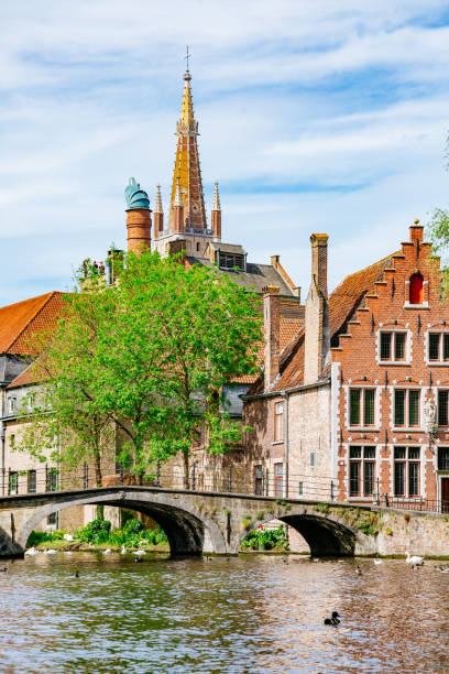 View of a canal in Bruges, Belgium. The canal is lined with historic brick buildings and a stone bridge. In the background, a church spire can be seen rising above the rooftops. The photo was taken on a sunny day, with blue skies and green trees visible.