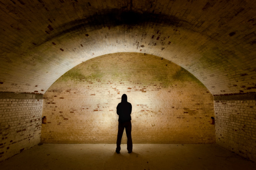 A shadowy figure in an underground tunnel faces a brick wall that is illuminated by a light in front of the figure.
