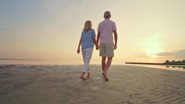 SLO MO TS Senior couple holding hands while walking barefoot on a sandy beach at sunset