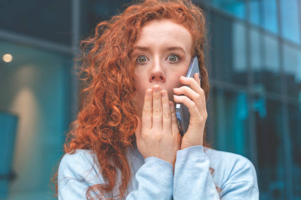 porträt einer schönen, schockierten jungen frau mit lockigem rotem haar, die auf dem handy spricht, während sie schreckliche nachrichten erhält - vacancy englisches schild stock-fotos und bilder