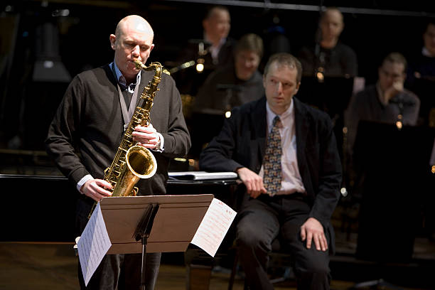 bigband: sax solo A solo saxophonist plays on stage while the musical director and rest of the band listen in the background. big band jazz stock pictures, royalty-free photos & images