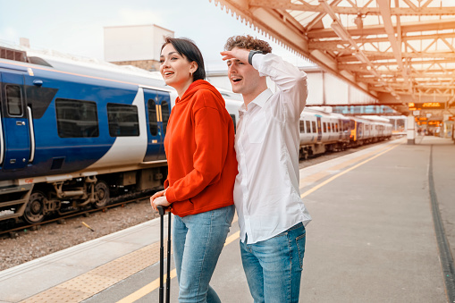 Happy young couple in excitement waiting for train at a railway station platform to go on holiday together