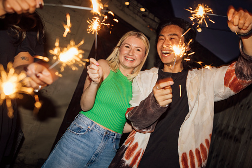 A young couple are at a New Year's Eve house party, they are nicely dressed and standing side by side in the garden holding sparklers on a dark night. They are celebrating the New Year in style with friends.