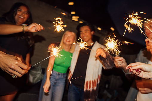 A small group of friends are at a New Year's Eve house party, they are nicely dressed and standing in a circle in the garden holding sparklers on a dark night. They are celebrating the New Year in style.